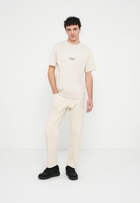 Beige cotton t-shirt with short sleeves and logo, paired with light beige trousers. Model wears black sneakers. Minimalist design.