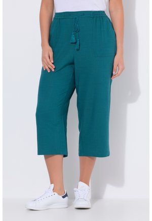 Pantalon large court couleur sarcelle avec taille à cordon et poche avant, porté avec des baskets blanches et un haut blanc.