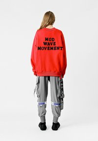 Sudadera roja con "MOVIMIENTO MOD WAVE" en letras negras en la parte posterior, combinada con pantalones cargo grises y calzado negro.