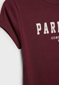 Burgundská bavlněná tričko s krátkým rukávem, které má bílý potisk s textem "PARIS" a "CONCEPT." Měkká textura a detaily s kruhovým výstřihem.