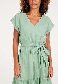 Hellgrünes Kleid aus strukturiertem Stoff, mit V-Ausschnitt, kurzen Ärmeln, einem Stoffgürtel und einem schichtweisen Rockdesign.