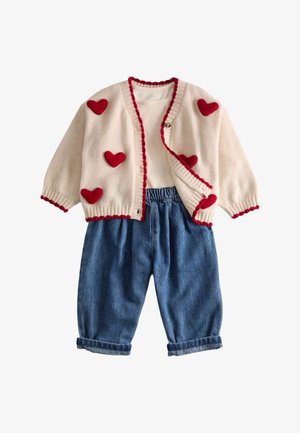 Cremefarvet cardigan med røde hjerte patches og kant, kombineret med hvid langærmet skjorte og blå denimbukser med opsmøg til et barn.