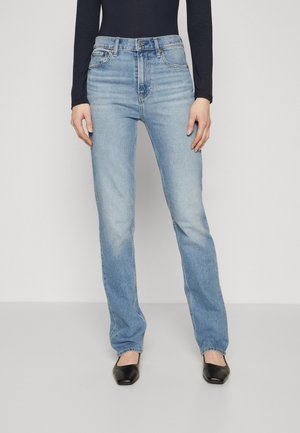 Femme portant un jean bleu clair droit, un haut noir à manches longues et des talons bas noirs à bout pointu, debout devant un fond uni.