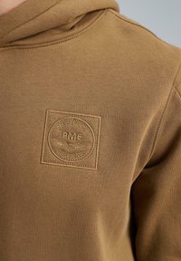 Brown hoodie featuring a textured patch with embossed logo. Smooth fabric with a relaxed fit and visible stitching on the edges.