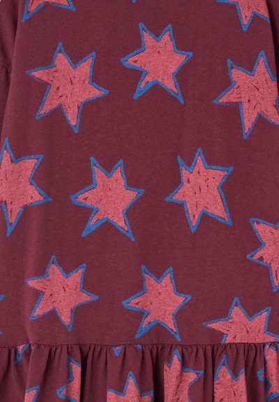 Burgunderfarbener Stoff mit einem Muster aus pinken Sternen, die blau umrandet sind. Die Sterne variieren in Größe und Unregelmäßigkeit, mit einem gerafften Saumdetail am unteren Ende.