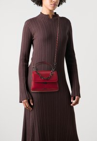 Raudona odinė rankinė su viršutine laikikliu ir grandinine juosta, pasižyminti lygiu paviršiumi ir uždarymu su užsegimu. Nešiojama kartu su rausvu ripsiniu suknele.