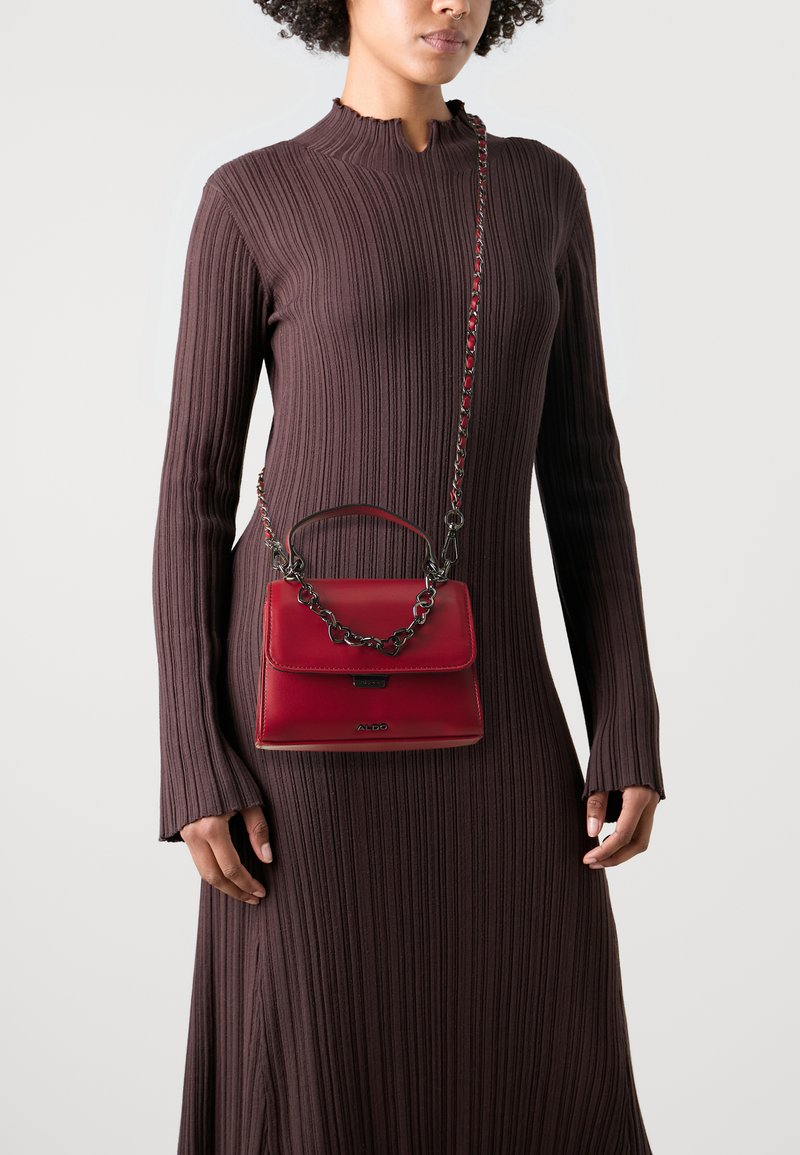 Raudona odinė rankinė su viršutine laikikliu ir grandinine juosta, pasižyminti lygiu paviršiumi ir uždarymu su užsegimu. Nešiojama kartu su rausvu ripsiniu suknele.