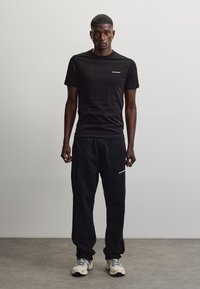 Mand står med fronten frem iført en sort t-shirt, sorte træningsbukser og hvide sneakers i en enkel indendørs setting.