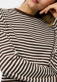 Osoba u smešnoj i beloj horizontalno prugastoj majici dugih rukava, s jednim rukama prekriženim i rukom oslonjenom blizu lica.