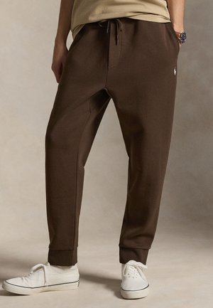 Mężczyzna w brązowych spodniach dresowych z troczkiem i białych trampkach, stojący ze złożonymi rękami w kieszeniach na neutralnym tle.