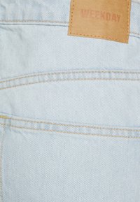 Tecido de denim azul claro com uma textura suave, apresentando uma etiqueta de couro castanho rotulada "WEEKDAY" e detalhes em costura amarela.