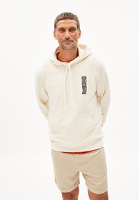 Felpa crema con tasca a marsupio frontale e un design grafico nero sul lato sinistro. Abbinata a pantaloni beige, presentata su uno sfondo bianco.