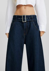 Mörkblå vida jeans med en tjock midja, prydda med en rektangulär silverfärgad bältesspänne och synliga sömnadsdetaljer.
