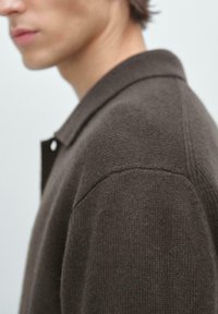 Boční pohled na osobu v tmavě hnědé pletené svetr s límcem na jednobarevném světlém pozadí.