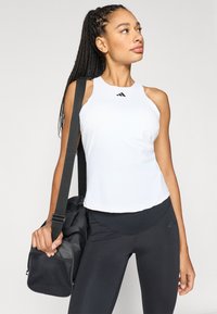 Camiseta deportiva blanca sin mangas con cuello redondo y logo negro, combinada con leggings negros. Modelo sosteniendo una bolsa de gimnasio negra con un panel de malla.