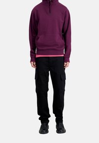 Sweat à capuche bordeaux avec une poche frontale et des cordons de serrage, superposé sur un t-shirt rose ; associé à un pantalon cargo noir et des chaussures noires.