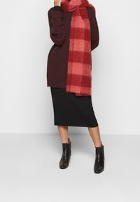 Pull en maille bordeaux, écharpe à carreaux rouges, jupe midi noire et bottines noires. Textures douces et couleurs contrastées.