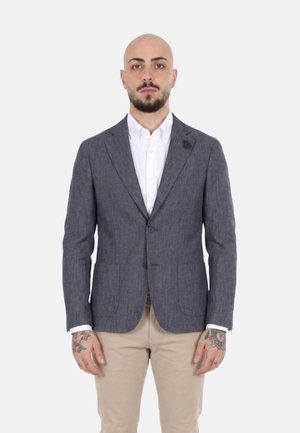 Uomo con tatuaggi sulle mani che indossa un blazer grigio, una camicia bianca e pantaloni beige, in piedi davanti a uno sfondo bianco semplice.