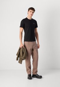 Tricou negru cu mâneci scurte și un logo mic, purtat cu pantaloni bronz deschis și pantofi negri masivi. O jachetă verde este aruncată pe un braț.