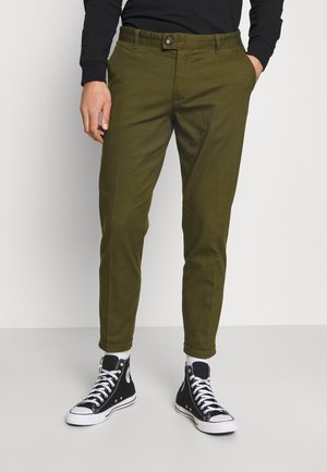 Man draagt olijfgroene slim-fit broek met opgerolde pijpen, zwart shirt met lange mouwen, witte sokken en zwarte hoge sneakers.