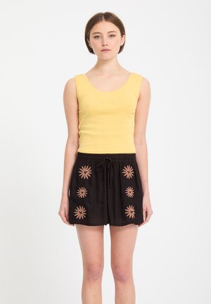 Jeune femme portant un haut jaune sans manches et un short noir avec six motifs floraux marron clair, debout devant un fond blanc uni.