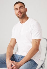Witte korte mouwen Henley-shirt met drieknoopsluiting, zachte katoenen textuur, gecombineerd met lichtblauwe jeans. Model zit op een stoel.