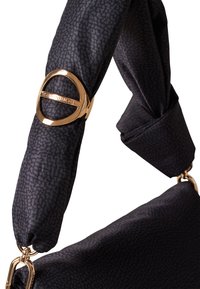 Sac à main en tissu noir avec une bandoulière nouée, présentant un détail en métal doré de forme circulaire. Le matériau a une texture et un motif subtils.
