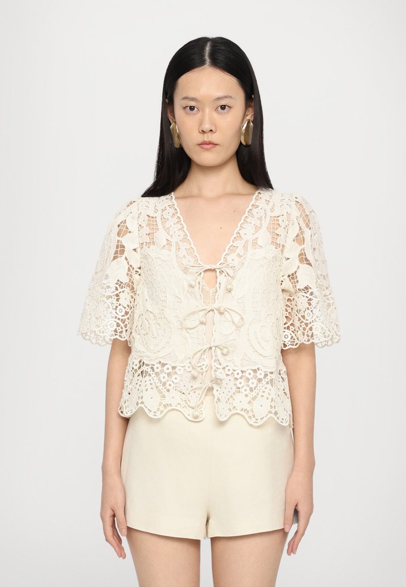 Valgetu lace blouse lühikeste hõljuvate varrukatega, lainelise ääre ja sügava V-kaelusega, millel on eesstringiga side.