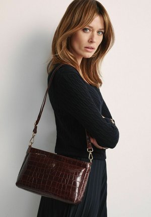Kobieta o jasnobrązowych włosach, ubrana w czarny sweter i spódnicę, niosąca ciemnobrązową, teksturowaną torebkę na ramię ze złotymi elementami.