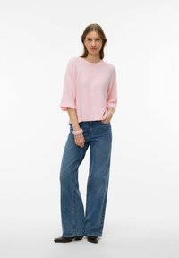 Svetlo roza pleten pulover s širokimi rokavi do komolcev, kombiniran z visokim pasom, ohlapnimi modrimi kavbojkami in temnimi gležnjarji z visokimi petami.