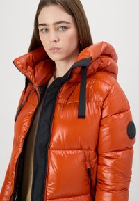 Bright orange puffer jacket with a shiny finish, featuring a black zipper, detachable hood, and black accents. Smooth texture with quilted design.