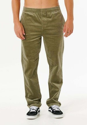 Persona con pantalones de pana verde oliva sueltos y zapatillas blancas y negras, de pie contra un fondo claro y liso.