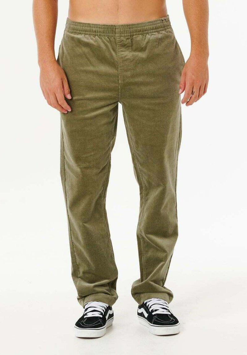 Person iført løse olivengrønne manchesterbukser og sort-hvide sneakers, stående mod en ensartet lys baggrund.