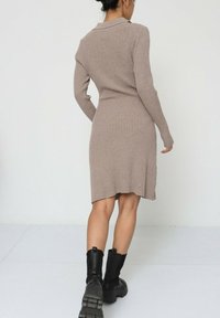 Robe pull en taupe, avec une texture côtelée, des manches longues et un ourlet légèrement asymétrique. Associée à des bottes épaisses noires à la cheville.