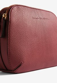 Primo piano di una pochette in pelle rosa scura texturizzata con zip dorata e piccolo dettaglio di cucitura nera su sfondo bianco.