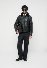 Veste en cuir noir avec une finition brillante et un col en fausse fourrure, associée à un pantalon noir et des chaussures noires. La tenue est accessoirisée avec un sac noir.