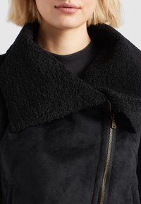 Femme portant une veste en peau lainée noire avec un grand col texturé et une fermeture éclair en laiton sur le côté droit devant.