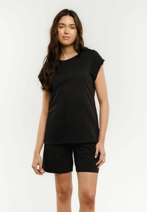 Mladá žena stojí oblečená v jednoduchom čiernom tričku s krátkym rukávom a čiernych krátkych nohaviciach na svetlom pozadí.