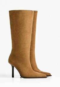 Bottes en daim hauteur genou de couleur marron clair avec un bout pointu et un talon aiguille. Texture lisse, coutures minimales sur les côtés, aucun matériel visible.