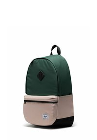 Sac à dos en tissu vert avec une section inférieure beige, étiquette noire triangulaire, poignée supérieure et poche avant zippée. Texture lisse et forme arrondie.