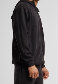 Sudadera negra con cremallera, fabricada en un tejido suave, que cuenta con una capucha forrada, bolsillos frontales y puños acanalados. Textura lisa con un corte relajado.