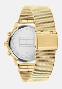 Montre en acier inoxydable doré avec un bracelet en maille, un boîtier rond et un verre transparent. L'arrière affiche le logo de la marque et les détails de résistance à l'eau.