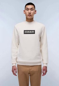 Giovane uomo in piedi, che indossa una felpa beige di Napapijri e pantaloni marrone chiaro, su sfondo neutro.