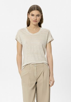 Frau mit langem braunem Haar, die ein beige-weiß gestreiftes T-Shirt mit kurzen Ärmeln und beige High-Waist-Hosen trägt, steht mit den Händen in den Taschen.