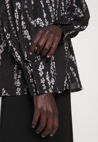 Mains à la peau foncée ajustant le poignet d'un chemisier noir à motif floral blanc avec des boutons recouverts de tissu, associé à un pantalon noir.