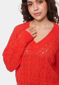 Donna con capelli ricci che indossa un maglione rosso a collo a V realizzato a maglia con un motivo in pizzo, mentre tiene il maglione vicino alla clavicola.