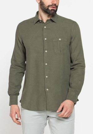 Uomo con la barba che indossa una camicia verde a maniche lunghe con taschino sul petto, abbinata a pantaloni grigio chiaro, in piedi su sfondo bianco.