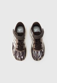 Pantofi sport cu un model camuflaj maro, având partea superioară din material textil, dungi albe, o curea maro cu Velcro și o talpă interioară gri texturată.