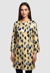Mujer vestida con un vestido de satén beige de mangas largas, impreso con pájaros azules y grises, de pie contra un fondo gris claro liso.