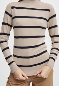 Maglione beige a collo alto con strisce orizzontali nere, polsini e orlo a costine. Texture morbida e design aderente, mostrato con le mani sull'orlo.